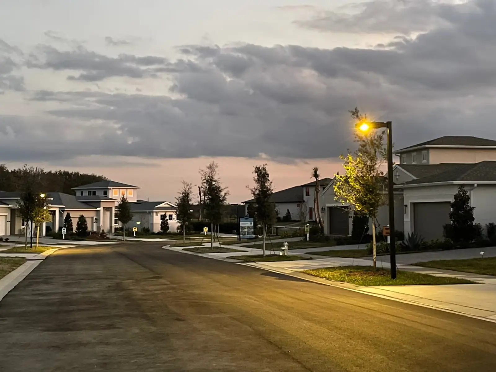 Residential street at dusk with modern houses, lit streetlamps, and partly cloudy sky.