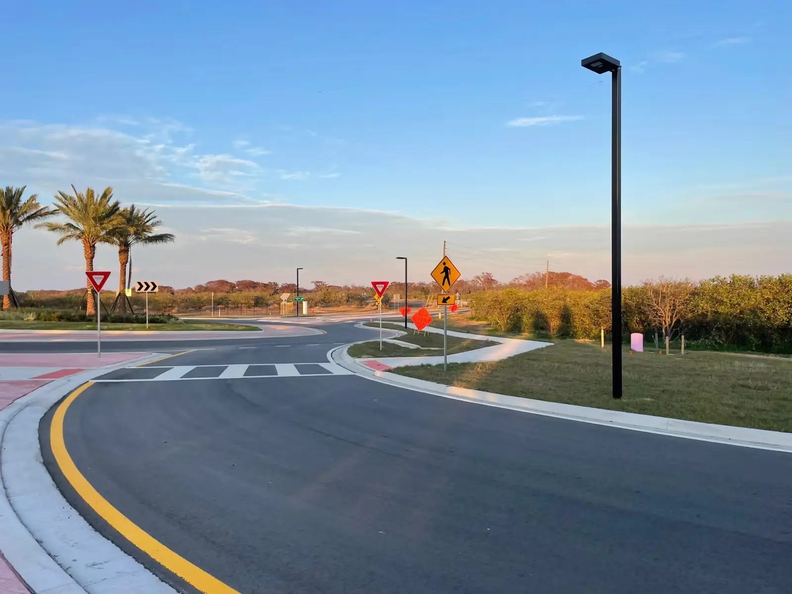 Empty curved road with crosswalk and multiple traffic signs, palm trees, streetlights, and clear sky at sunset.