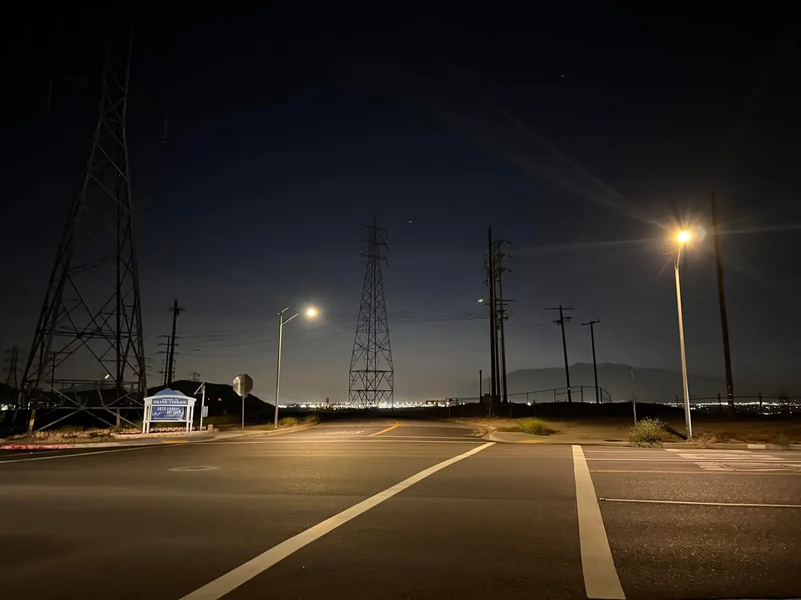 Empty street intersection at night with streetlights, power lines, and city lights in the distance.