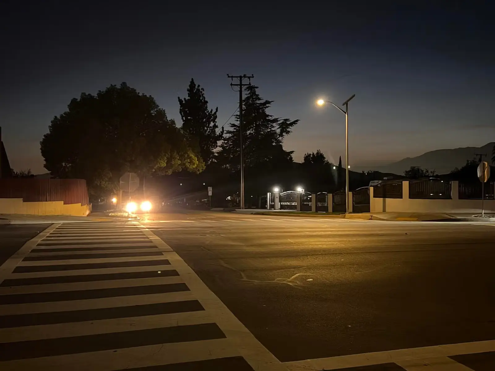 Nighttime street scene with a crosswalk, streetlight, trees, and car headlights shining toward the intersection.