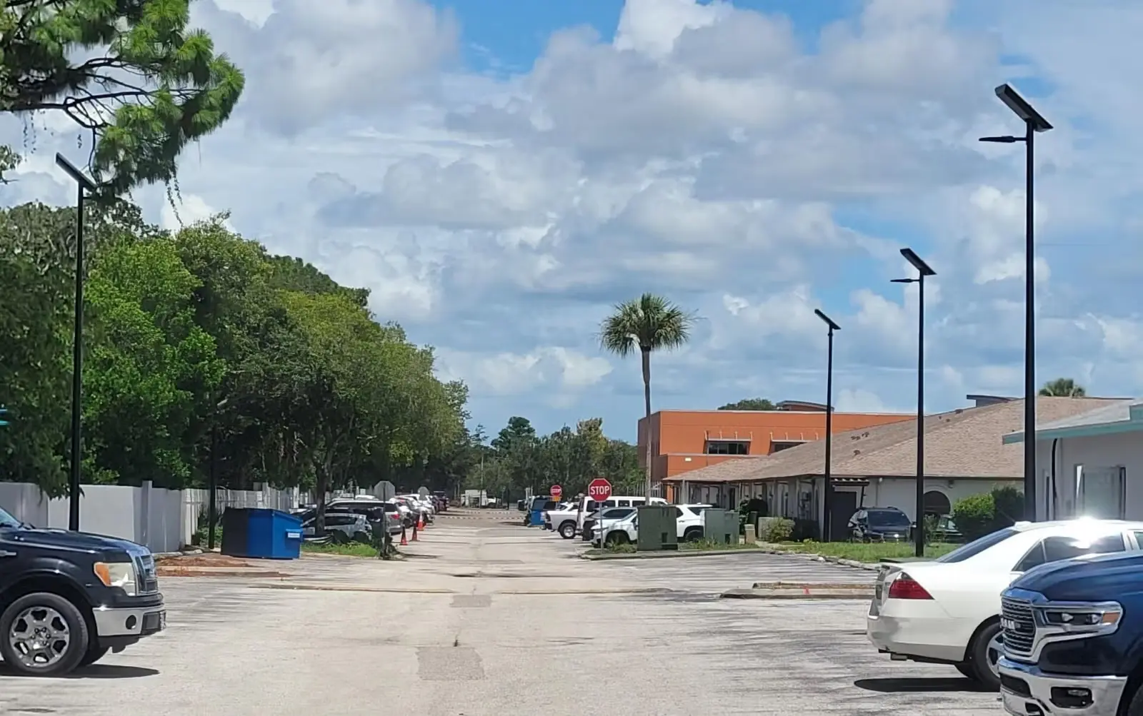 Parking lot with various parked cars, street lamps, trees, and a stop sign under a partly cloudy sky.