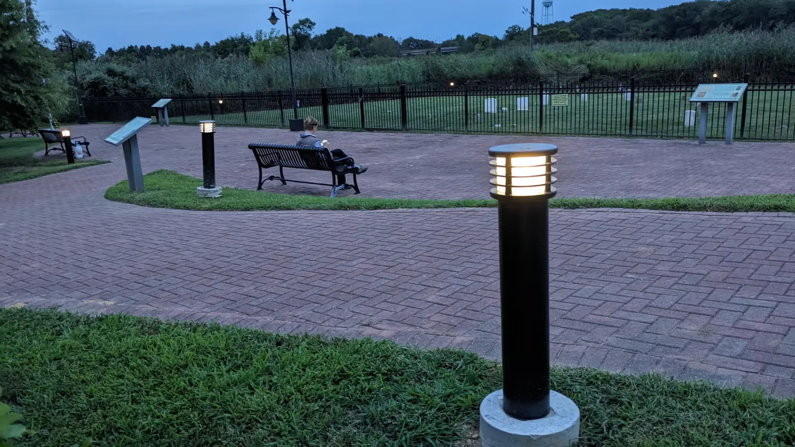 Person sitting on a bench in a park at dusk near illuminated path lights and brick-paved pathways.