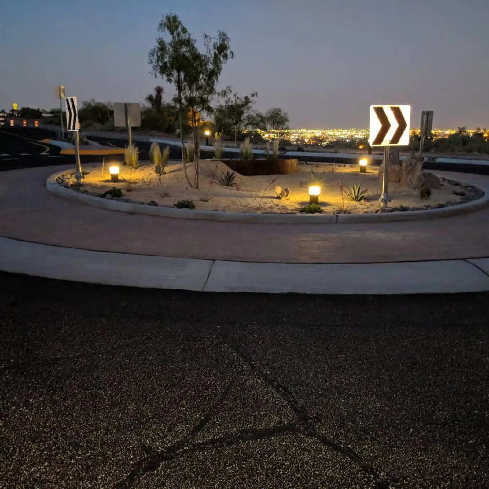 Night view of a desert-themed roundabout with illuminated arrow signs and city lights in the background.