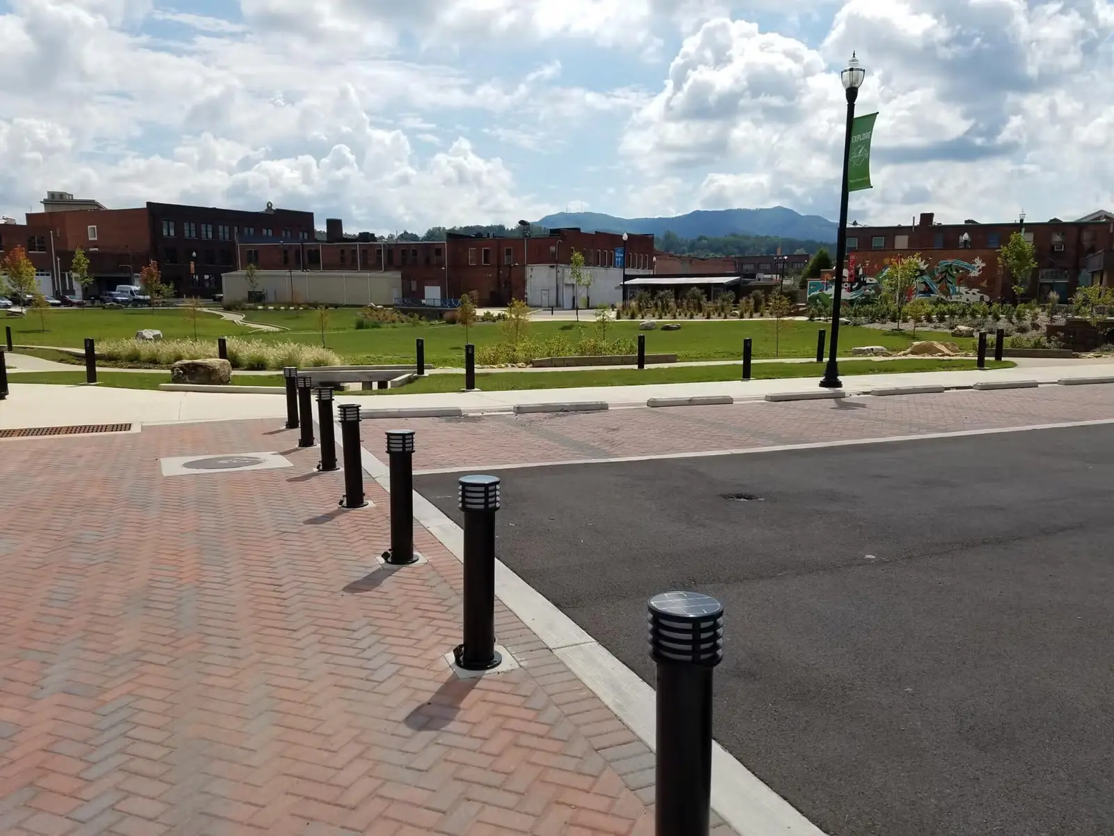 Urban park with green lawn, brick sidewalk, black bollards, street lamp, and red brick buildings under a partly cloudy sky.