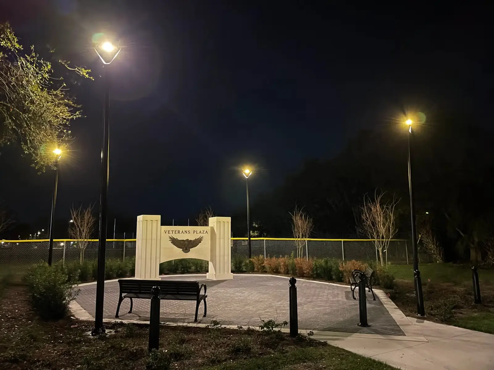 Night view of Veterans Plaza with illuminated lamp posts, benches, and a monument featuring an eagle emblem.