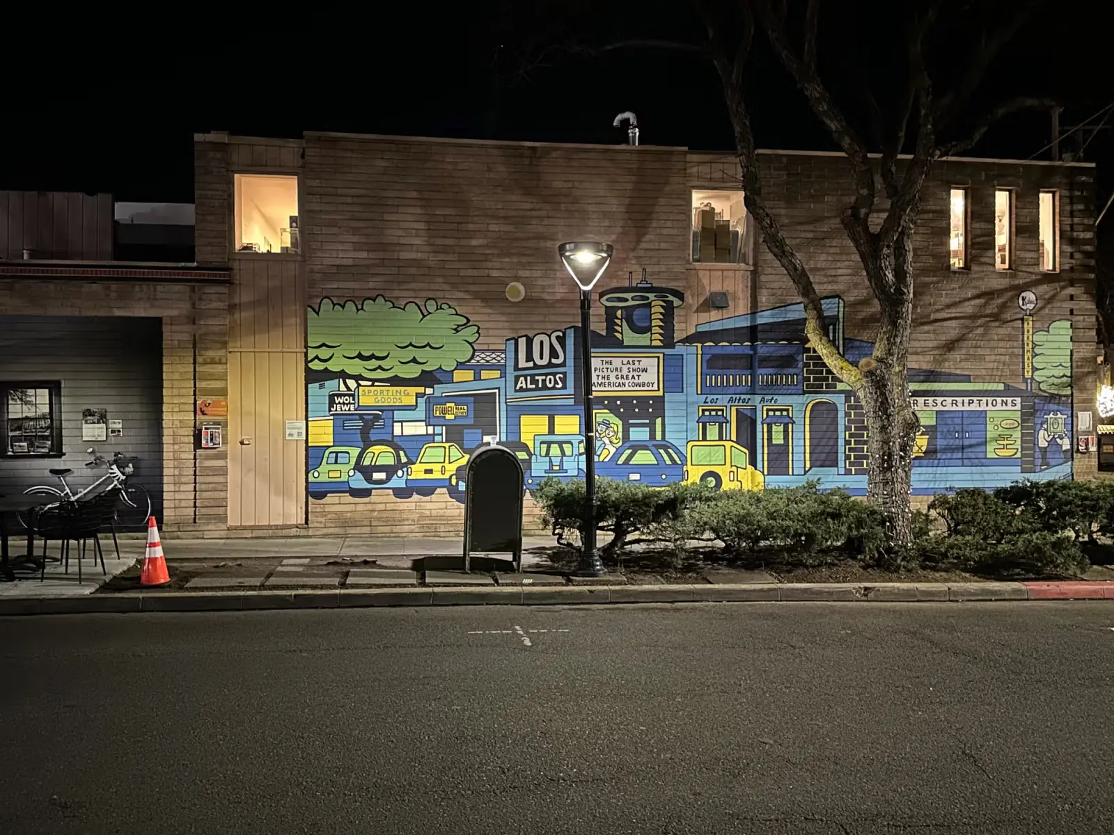 Street view at night featuring a building wall mural depicting a colorful cityscape with cars, trees, and storefront signs including 'Los Altos', 'Sporting Goods', and 'Prescriptions'.