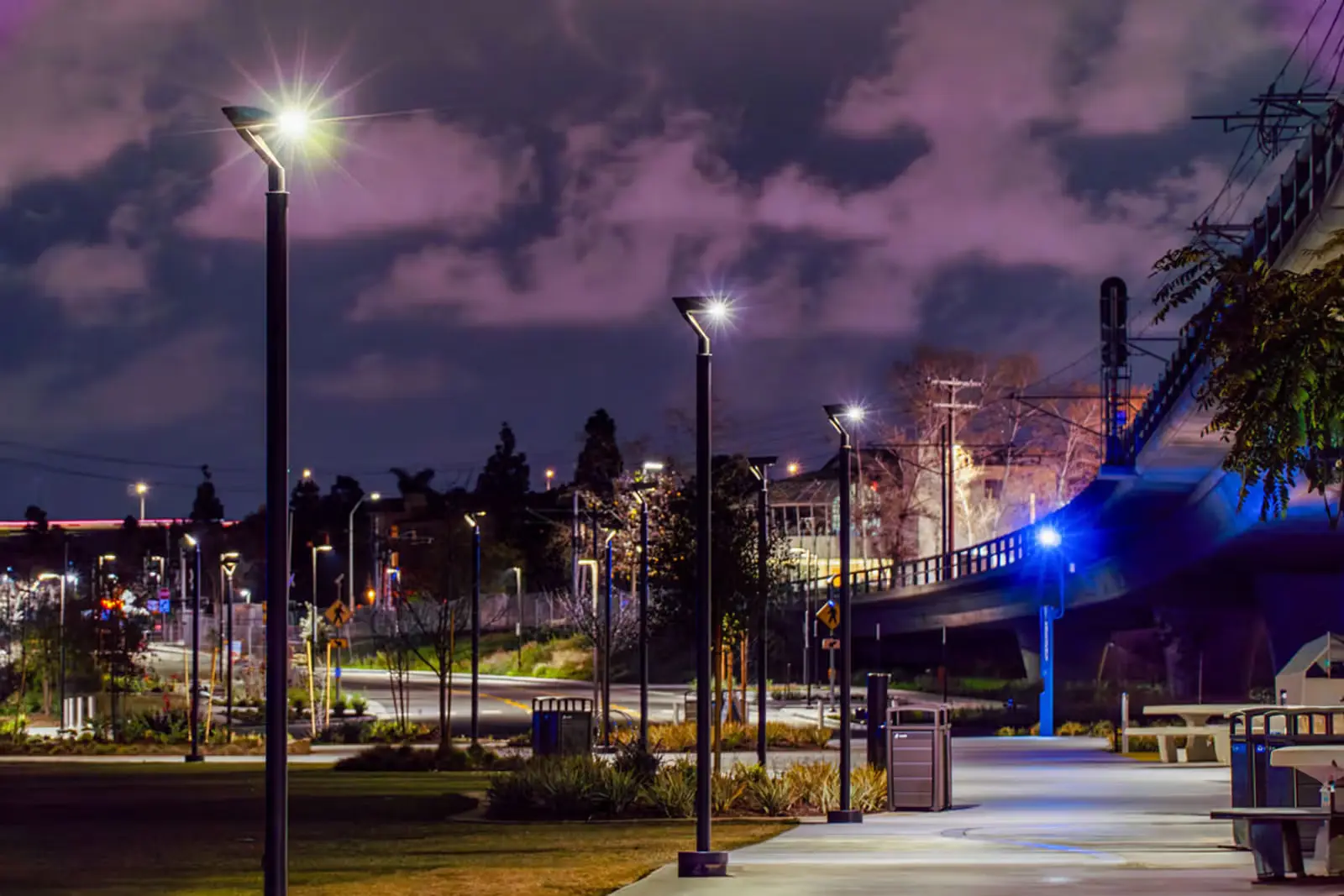 Night view of a modern park pathway illuminated by tall streetlights with a bridge and city lights in the background.