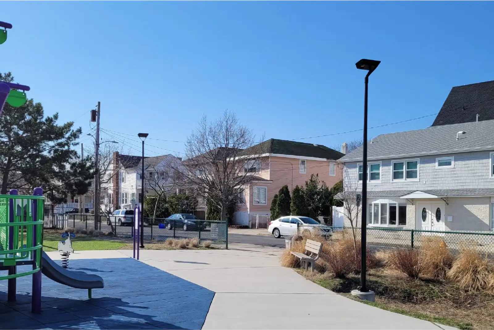 Playground area with a slide, bench, and residential houses in the background under a clear blue sky.