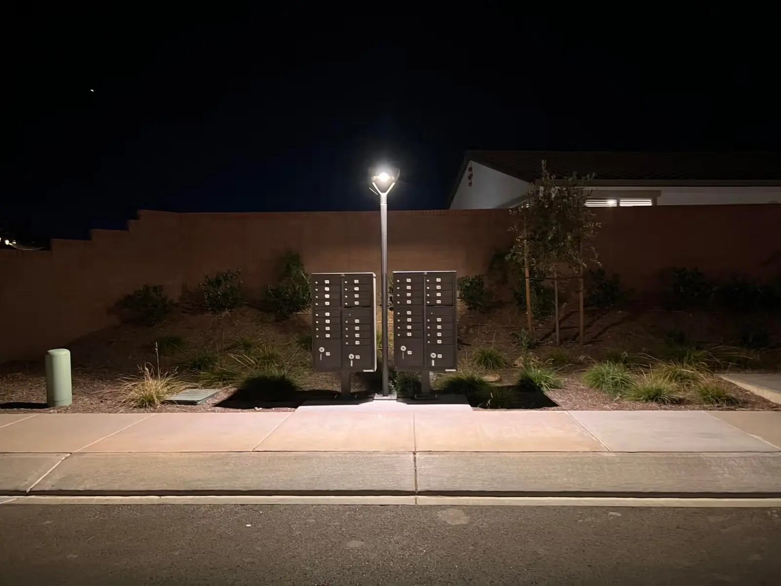 Two sets of black community mailboxes illuminated by a streetlamp at night, with a sidewalk and residential landscaping in the background.