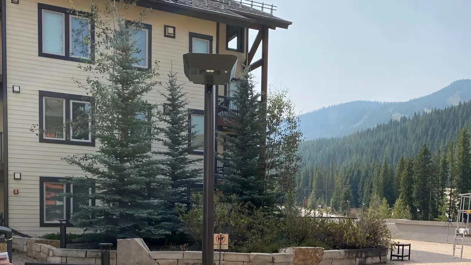 Outdoor area with spruce trees, a beige building with black window frames, stone wall planters, and distant forested mountains under a clear sky.