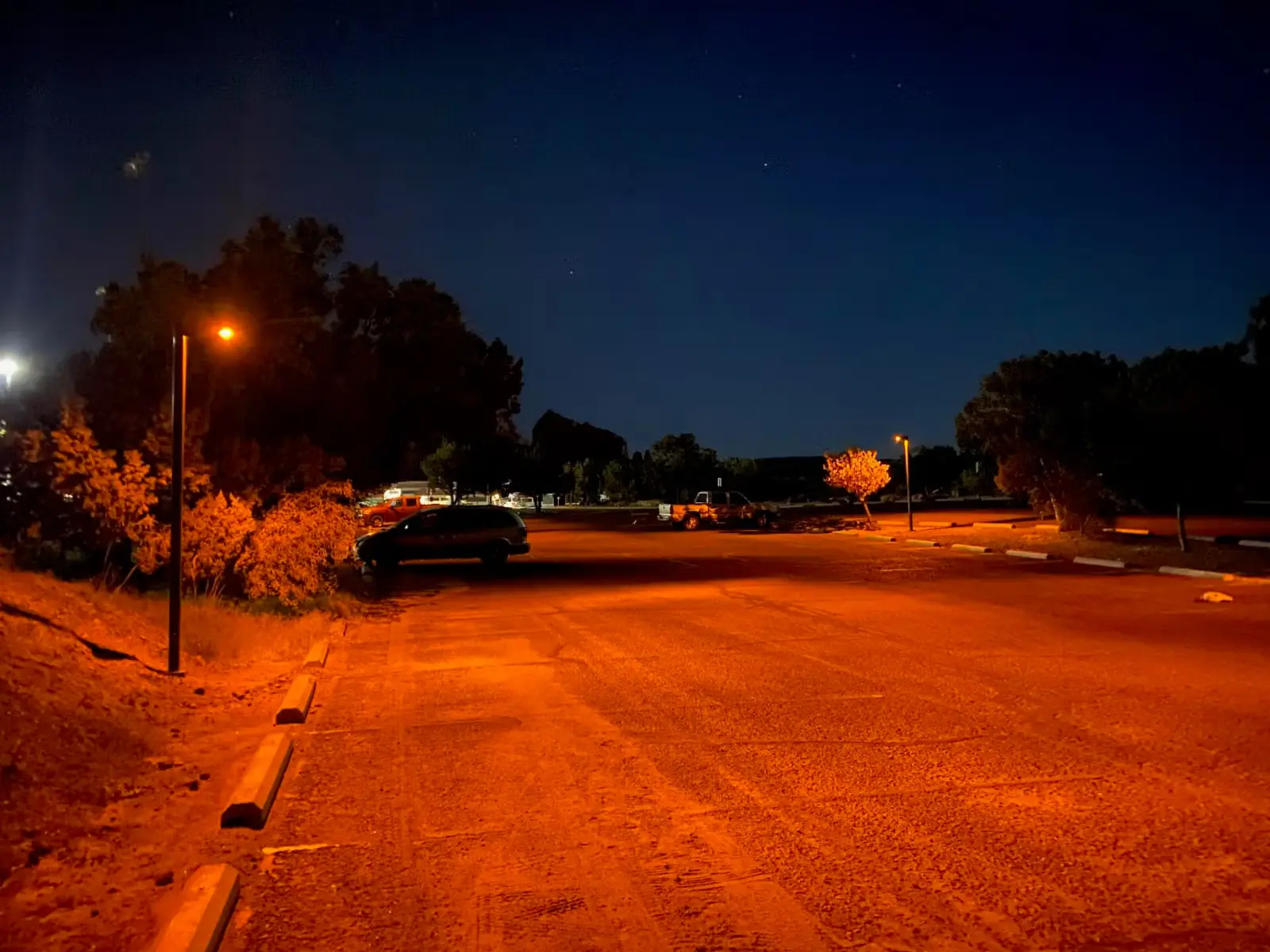 Quiet parking lot at night illuminated by orange streetlights with a few parked vehicles and trees in the background.