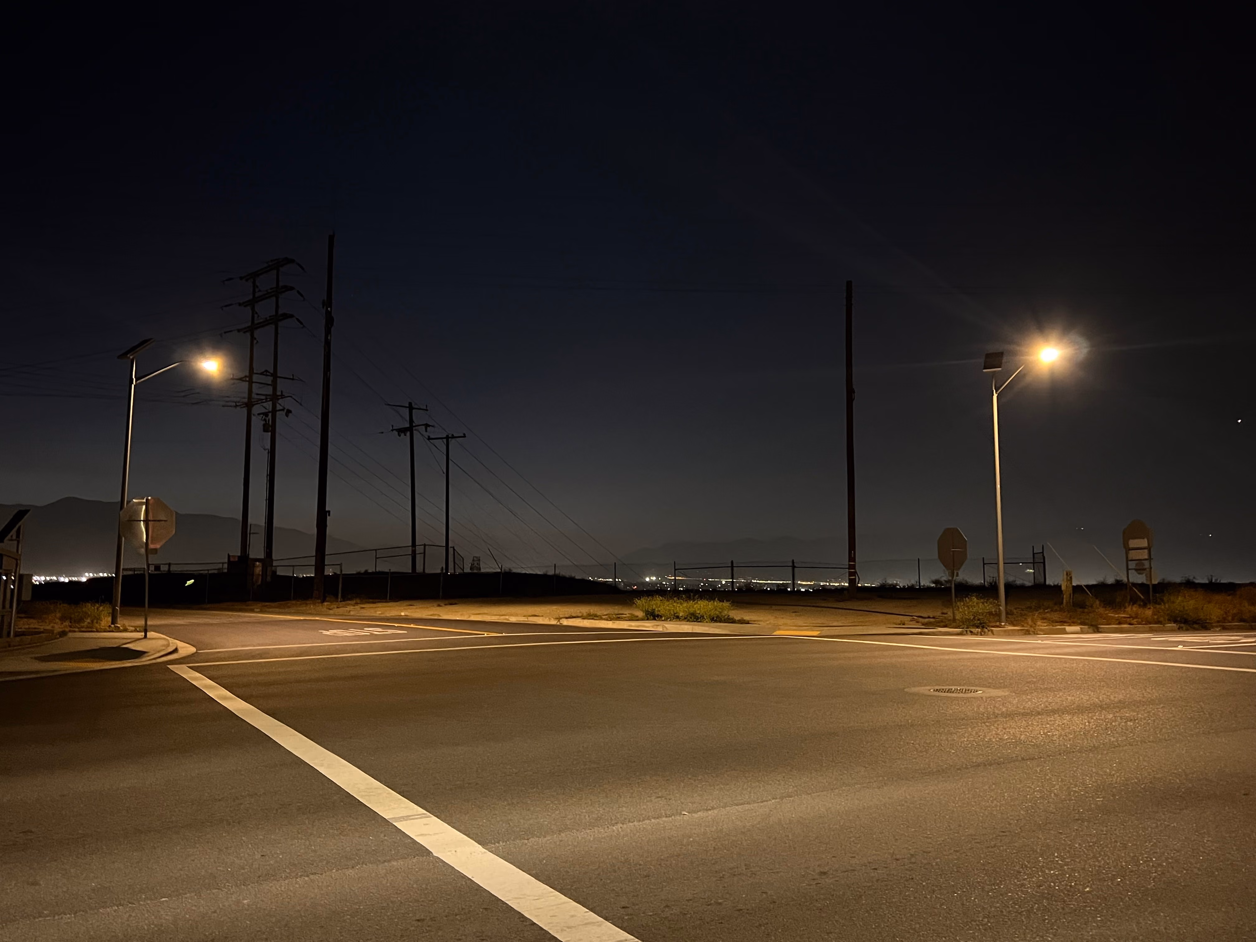Empty street intersection at night with streetlights and distant city lights and mountains.