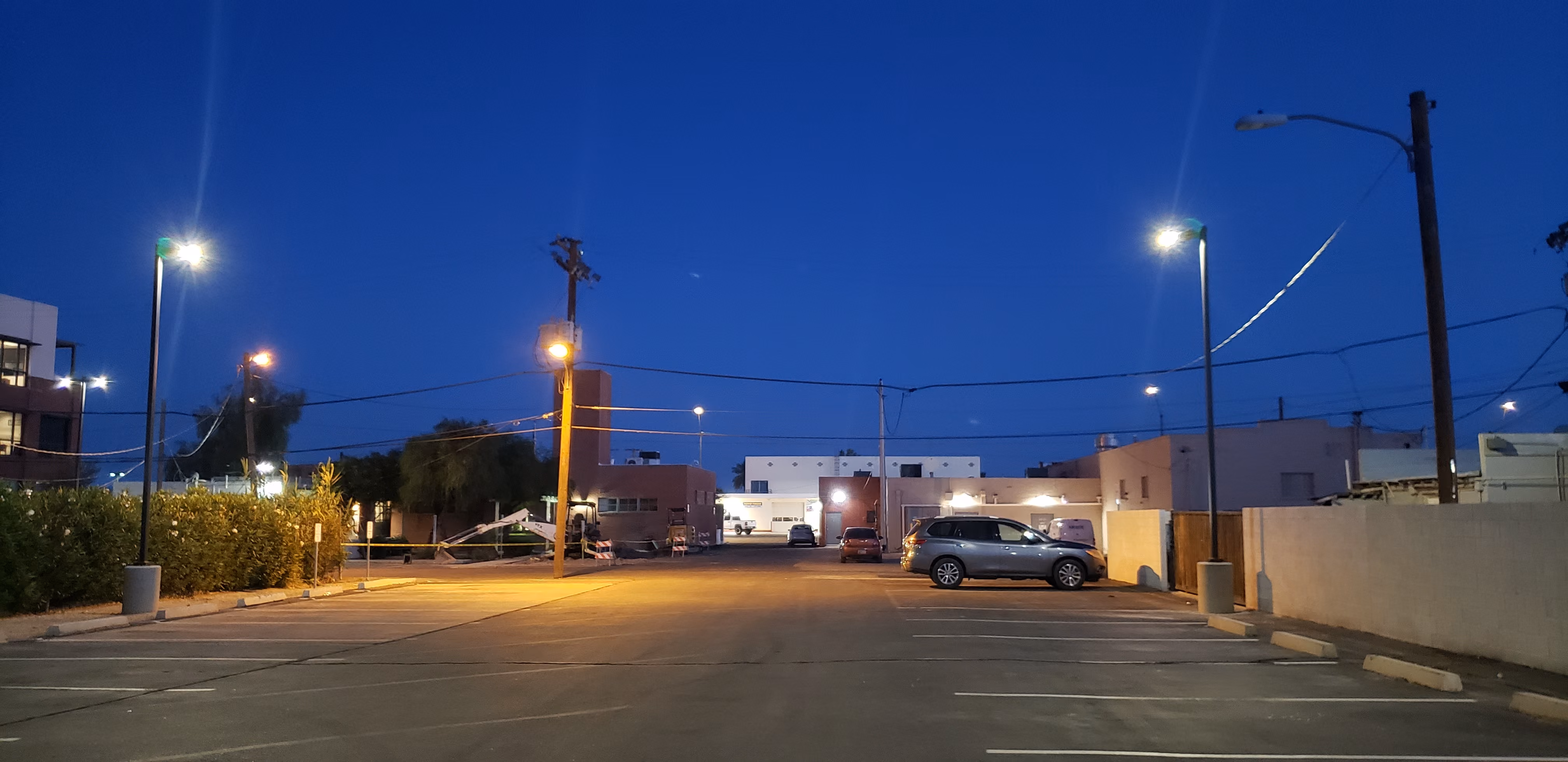Empty urban parking lot at dusk with illuminated street lamps, a few parked cars near beige buildings, and a dark blue sky overhead.