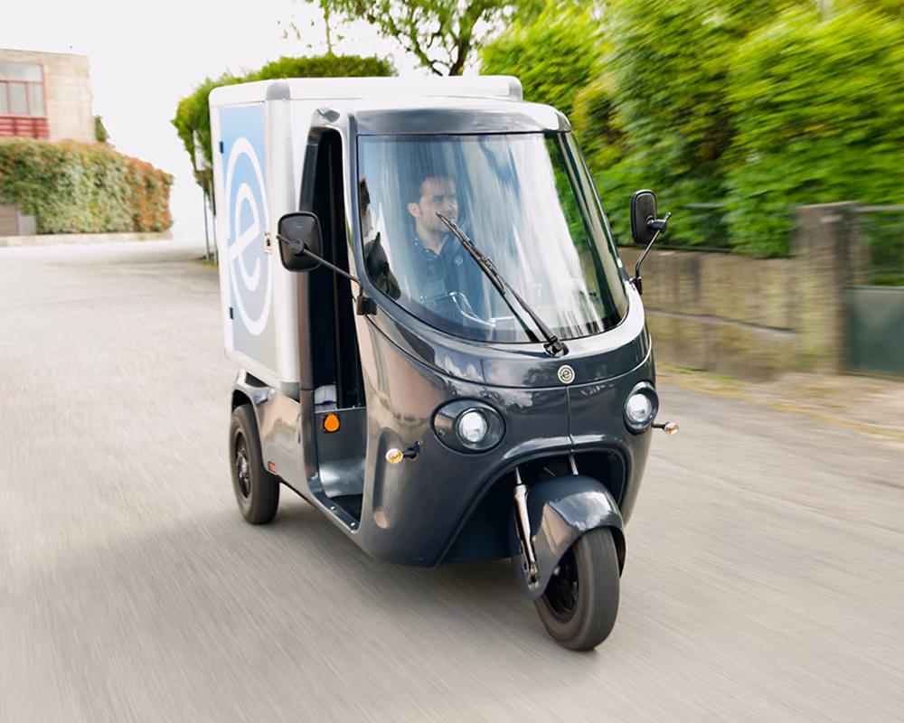 SmarTuk L2 mit Fahrer fährt auf einer Straße durch ein grünes Wohngebiet.