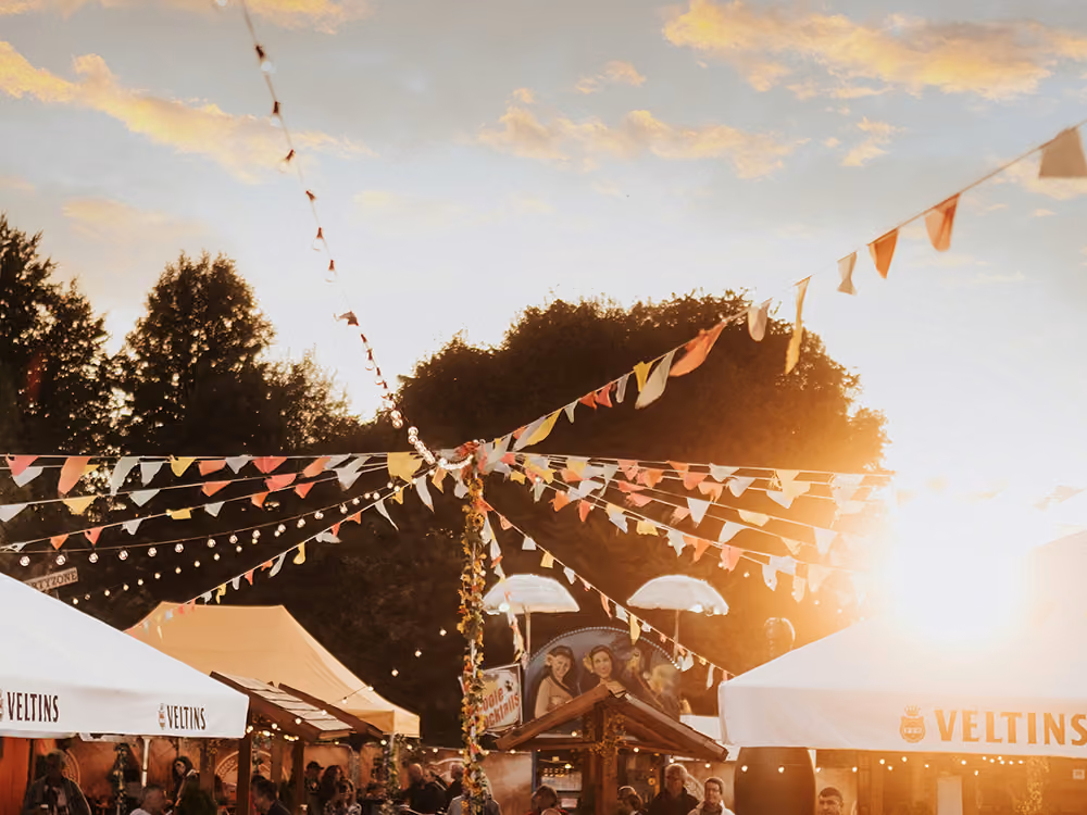 Sommerliches Festgelände bei Sonnenuntergang mit bunten Wimpelketten und Lichterketten über weißen Zelten, Marktständen und einer Menschenmenge.