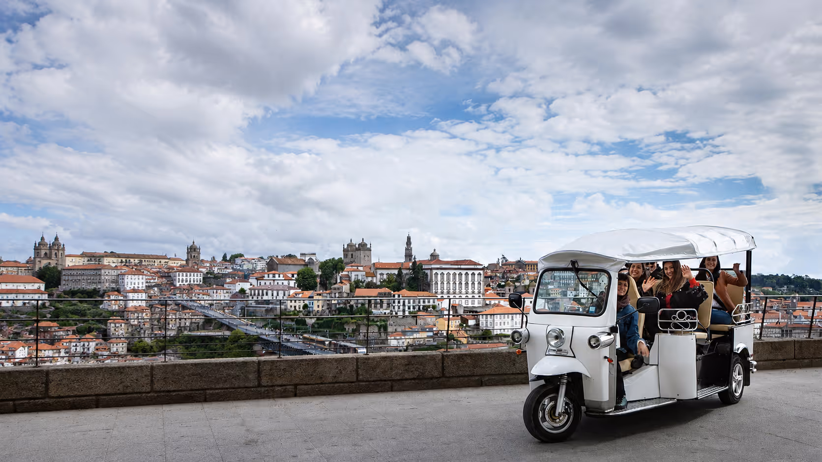 Eine Gruppe junger Frauen sitzt winkend in einem weißen L5 Limo GT auf einer Aussichtsplattform; im Hintergrund ist die Altstadt von Porto mit roten Dächern, Kirchen und bewölktem Himmel zu sehen.