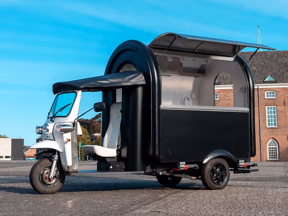 L5 eFoodTuk mit aufgeklappter Verkaufsklappe, geparkt auf einem gepflasterten Platz vor einem Backsteingebäude.
