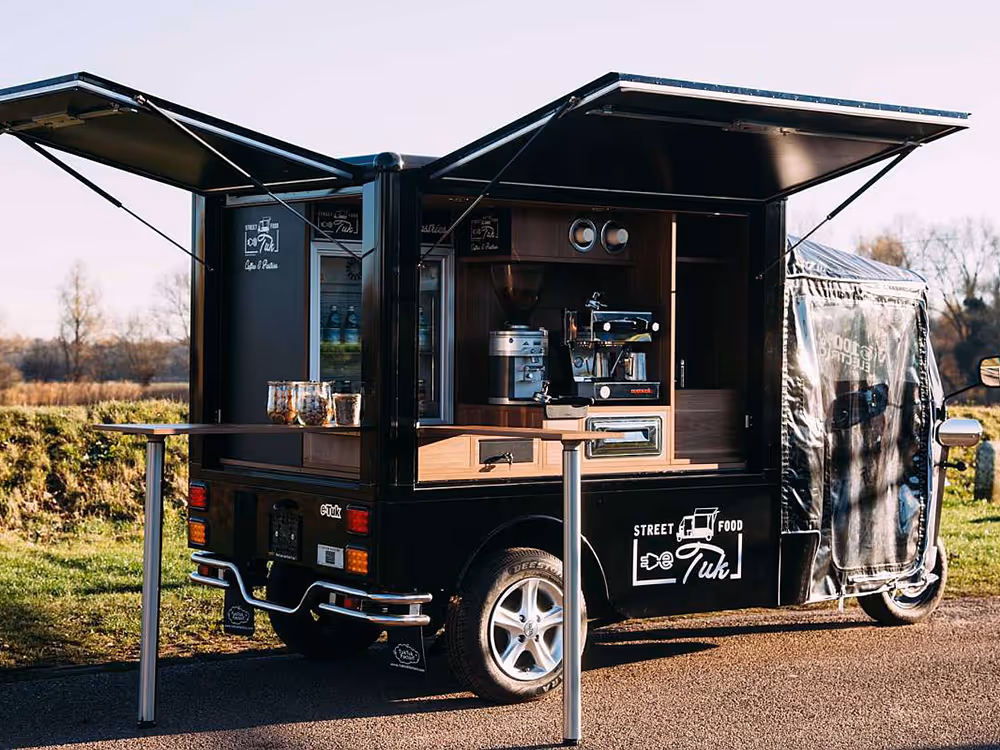 L5 Cargo mit aufgeklappten Verkaufsklappen, Espressomaschine und kleiner Theke, abgestellt auf einer Straße im Grünen.