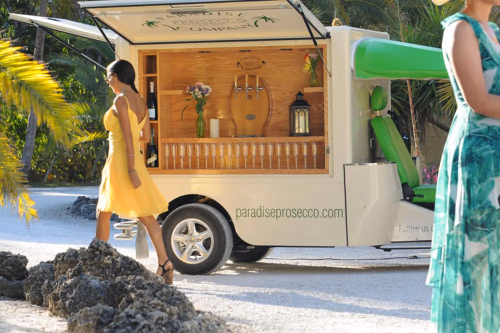 L5 Cargo als mobiler Prosecco-Wagen am Strand mit geöffneter Bar, tropischer Bepflanzung und zwei vorbeigehenden Frauen in Sommerkleidern.