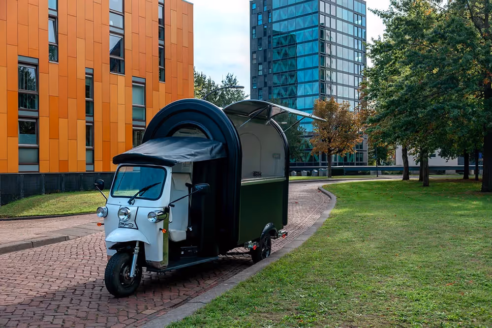 Ein L5 eFoodTuk steht auf einem gepflasterten Weg in einem modernen Stadtviertel mit Bürogebäuden, Grünfläche und Bäumen.