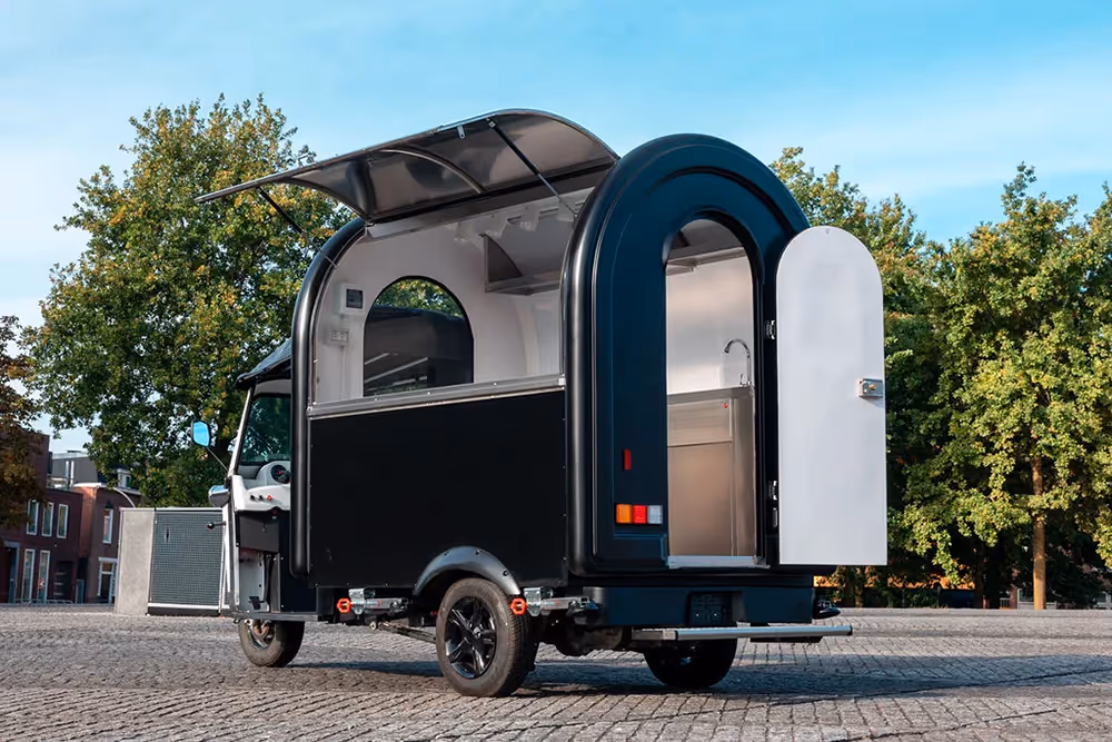 Schwarzer mobiler Foodtruck L5 eFoodTuk mit aufgeklapptem Verkaufsfenster und geöffneter Hecktür auf einem gepflasterten Platz, im Hintergrund grüne Bäume und Gebäude.