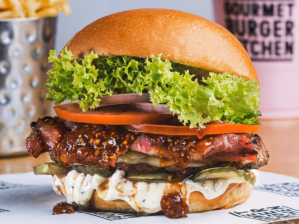 Großer Burger mit glänzendem Brioche-Brötchen, grünem Blattsalat, Tomaten, roten Zwiebeln, Gurken, cremiger Sauce und einem saftigen, glasierten Rindfleischpatty; im Hintergrund sind Pommes und ein Getränk zu sehen.