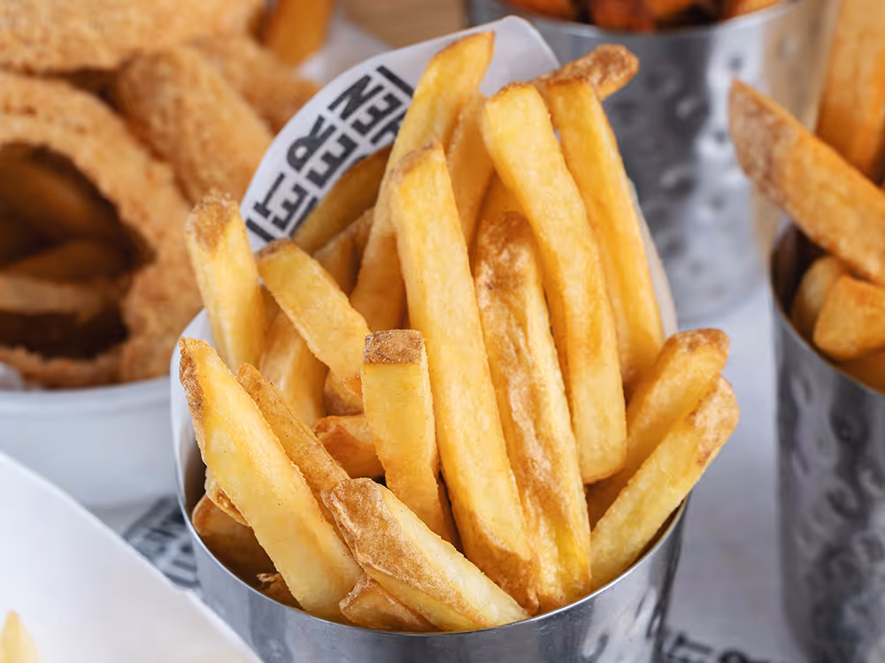 Goldbraune Pommes frites in einem Metallbecher, im Hintergrund unscharf weitere frittierte Beilagen.