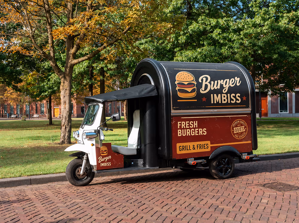 L5 eFoodTuk als Burger-Imbisswagen mit rot-schwarzem Aufbau und Beschriftung, geparkt auf einer Kopfsteinpflasterstraße in einem grünen Park.