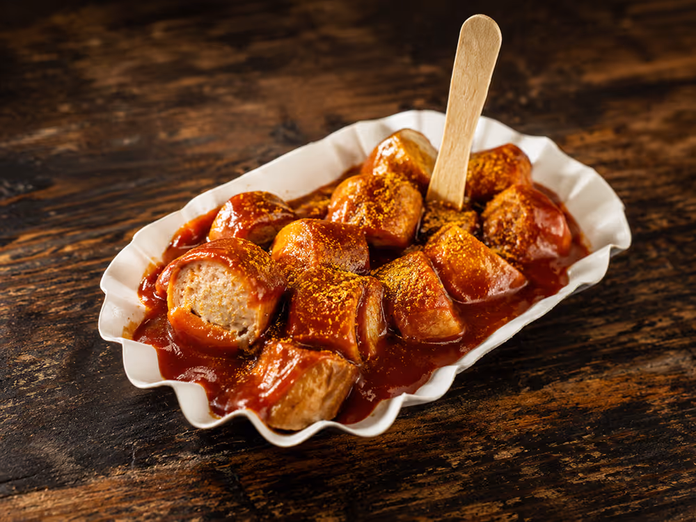 Currywurststücke mit roter Currysauce und Currypulver in einer weißen Pappschale, serviert mit einer kleinen Holzgabel auf einem rustikalen Holztisch.