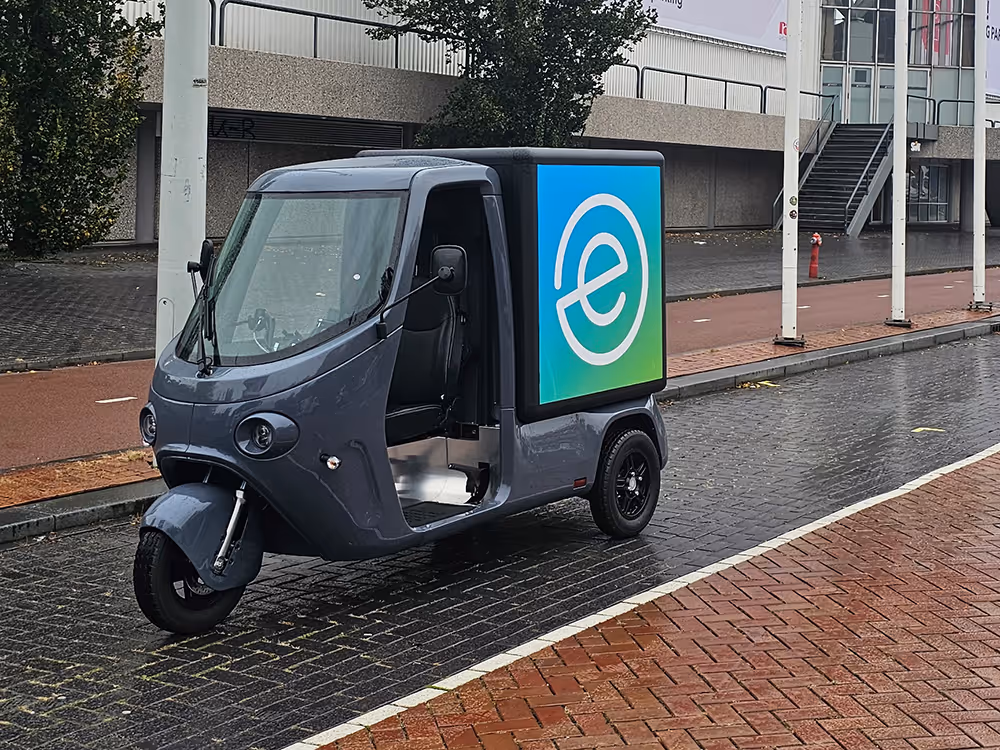 SmarTuk L2 mit großer Werbefläche an der Seite, geparkt auf einer nassen Stadtstraße neben einem Radweg.