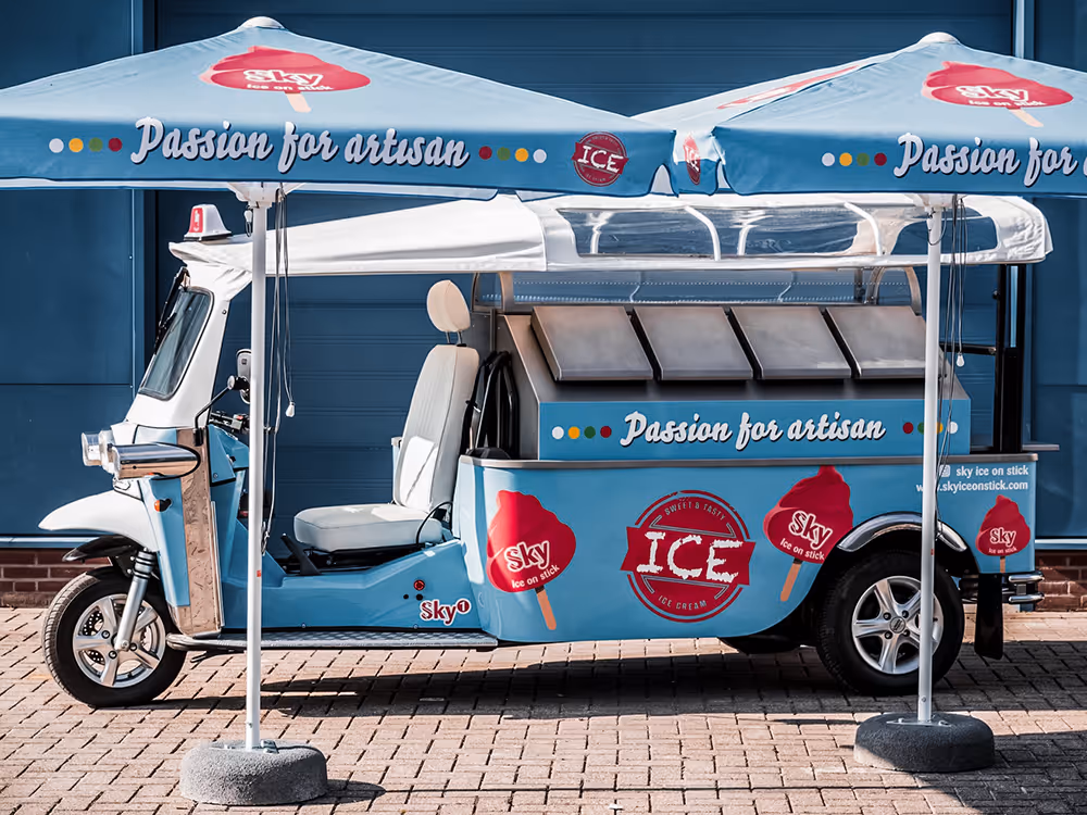 L5 Cargo in Hellblau mit Sky-Branding und der Aufschrift „Passion for artisan“, geparkt unter zwei passenden Sonnenschirmen vor einer blauen Gebäudewand.