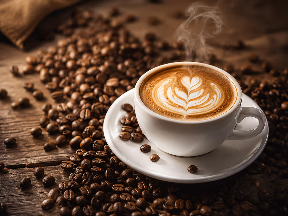 Weiße Tasse mit Latte Art auf einer Untertasse, umgeben von gerösteten Kaffeebohnen auf einem rustikalen Holztisch; aus dem Kaffee steigt Dampf auf.