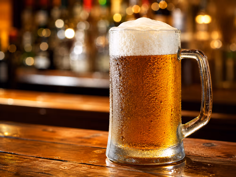 Ein großes, beschlagenes Bierglas mit goldenem Bier und dichter Schaumkrone steht auf einer hölzernen Bartheke vor unscharf beleuchtetem Hintergrund.