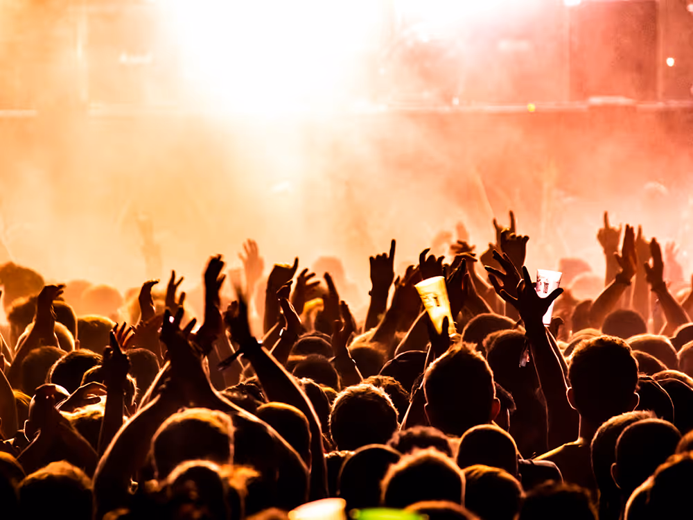 Menschenmenge bei einem Konzert mit erhobenen Händen und Getränkebechern vor einer hell orange beleuchteten Bühne.