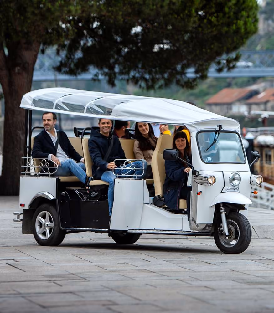 Eine Gruppe von fünf Personen fährt in einem offenen, weißen Elektro-Tuk-Tuk L5 Limo GT mit beigem Innenraum und weißem Stoffdach über einen gepflasterten Platz.