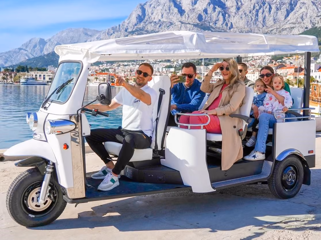 L5 Limo GT mit Fahrer und mehreren Passagieren steht an einer sonnigen Uferpromenade vor türkisblauem Wasser, einer Küstenstadt und steilen hellen Bergen im Hintergrund.