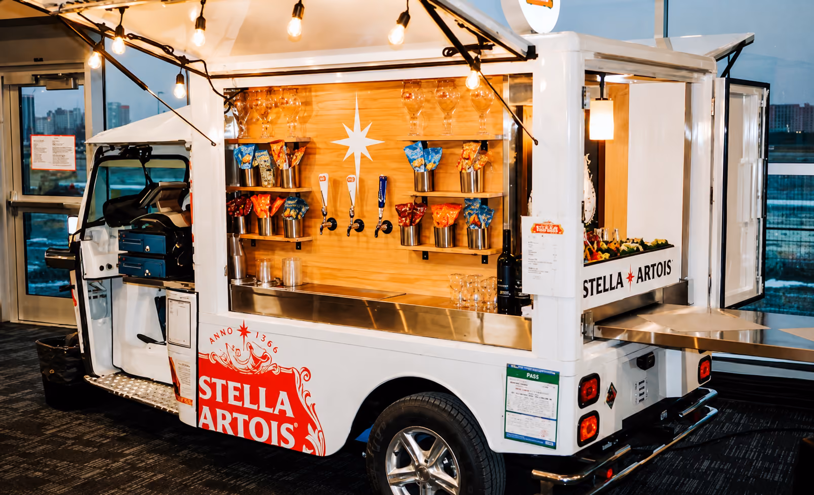 L5 Cargo als mobiler Stella-Artois-Ausschankwagen mit geöffneter Seitenklappe, Zapfhähnen, Becherhaltern und Lichterkette in einer Veranstaltungshalle.