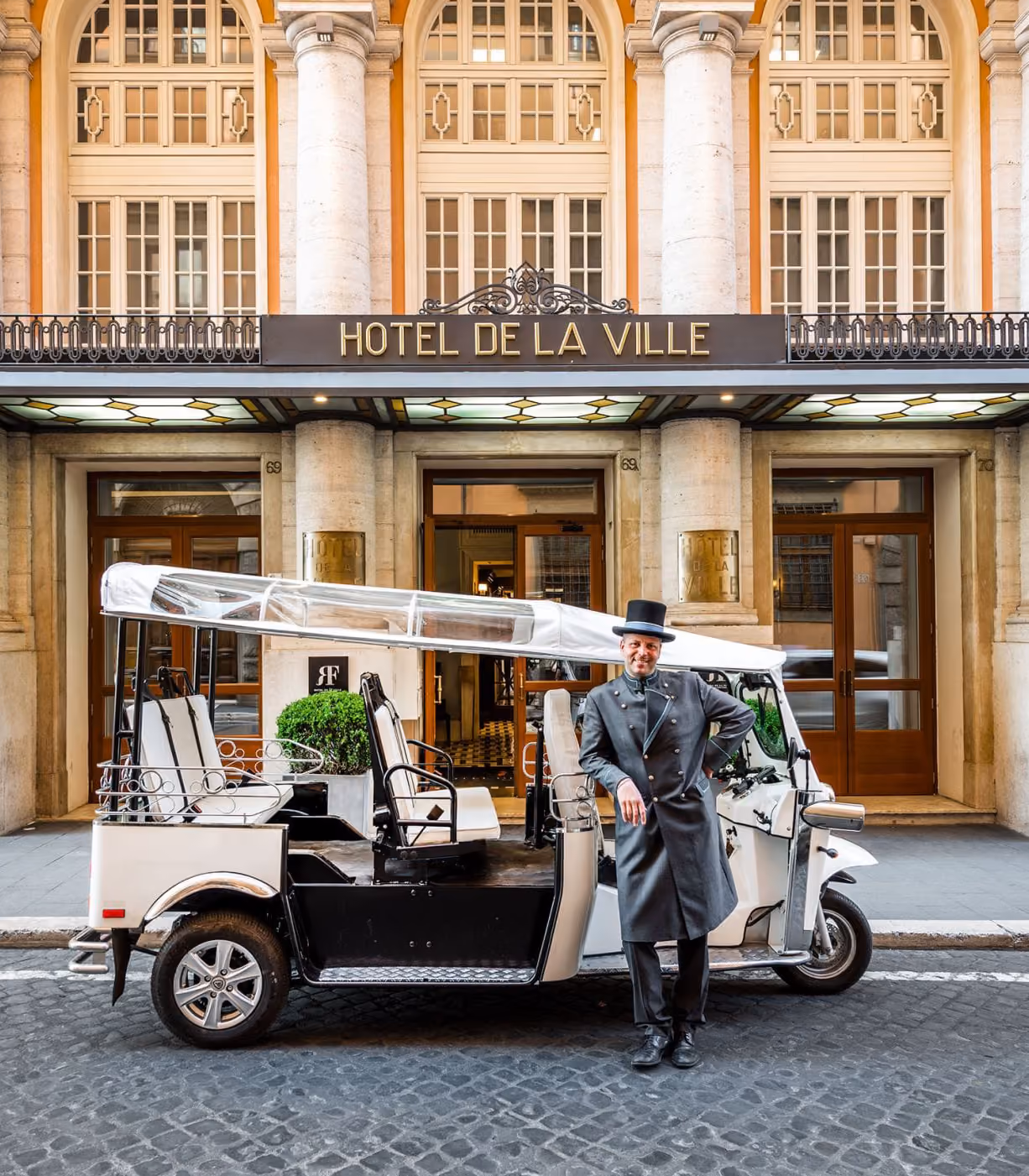 Eleganter Chauffeur in Livree steht vor einem weißen L5 Limo GT vor dem Eingang des Hotel de la Ville in einer historischen Stadtstraße.