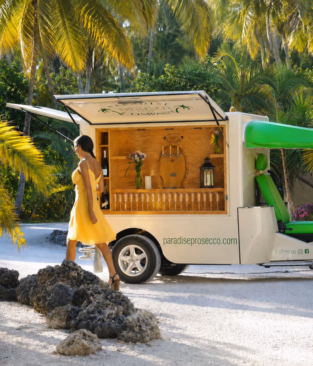 Eine Frau in einem gelben Sommerkleid geht an einem L5 Cargo als mobile Prosecco-Bar vorbei, die unter Palmen in einer tropischen Umgebung steht. Die weiße Bar ist geöffnet und mit Gläsern, Zapfhähnen, Blumen und einer Laterne ausgestattet.
