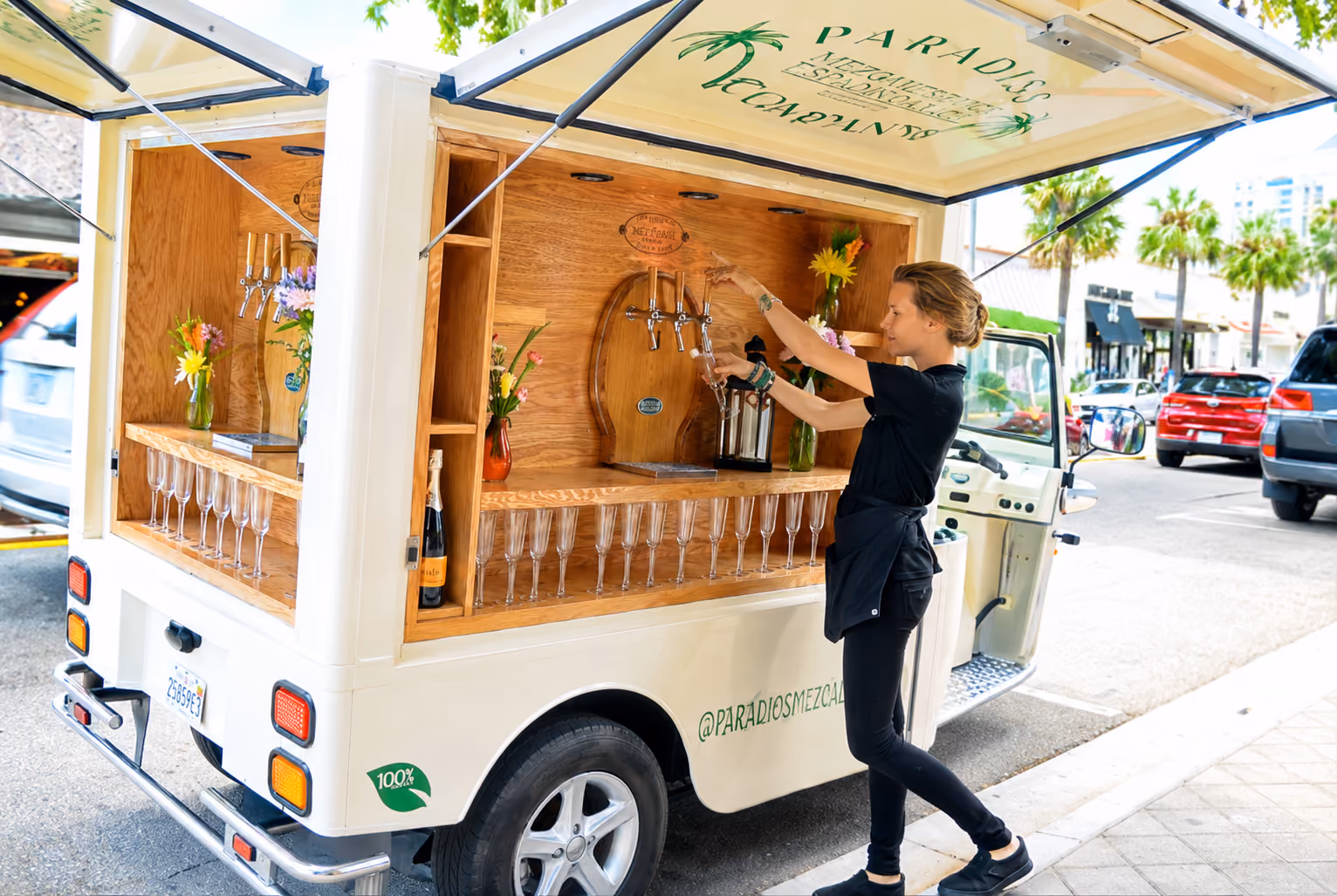 L5 Cargo als mobile Bar steht mit geöffneten Seitenklappen am Straßenrand. Eine Mitarbeiterin in schwarzer Kleidung zapft ein Getränk aus einem Holzfass; im Innenraum stehen zahlreiche Gläser und bunte Blumenvasen.