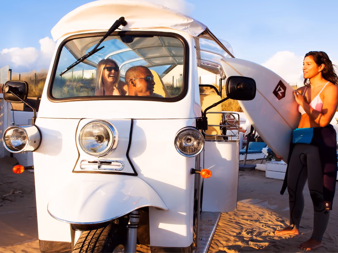 Drei Personen an einem Strand neben einem weißen Tuk-Tuk L5 Limo GT. Zwei sitzen lachend im Fahrzeug, während eine Frau im Neoprenanzug ein Surfbrett trägt und mit ihnen spricht.