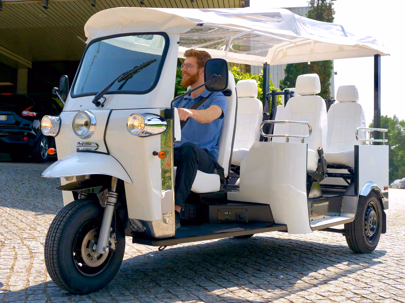 Weißes Elektro-Tuk-Tuk L5 Limo GT mit mehreren Sitzreihen und transparentem Dach. Ein bärtiger Mann mit Brille sitzt am Steuer; das Fahrzeug steht auf einem gepflasterten Platz vor der eTuk Factory.
