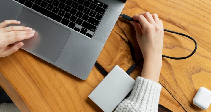 Person connecting an external hard drive to a laptop on a wooden table.
