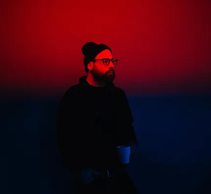 Man wearing glasses and a beanie holding a cup, illuminated by red light against a dark background.