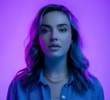 Portrait of a young woman with wavy hair wearing a blue shirt and layered gold necklaces against a purple-lit background.