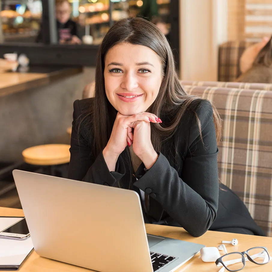Smiling young woman in a black blazer sitting at a table with a laptop, glasses, and earbuds in a cozy cafe.