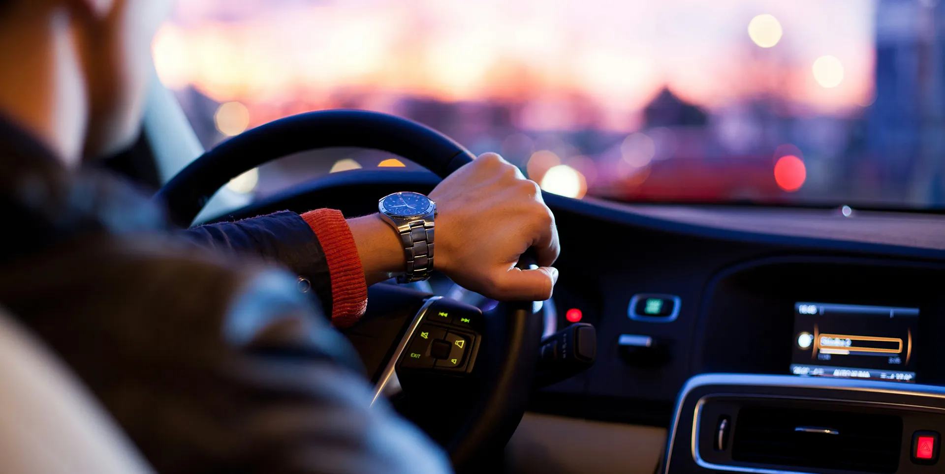 A driver's hand wearing a luxury watch on the steering wheel of a car at dusk