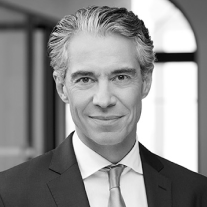 Smiling middle-aged man with gray hair wearing a suit and tie in a bright office setting.