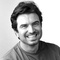 Smiling man with dark hair and stubble wearing a casual shirt against a white background.