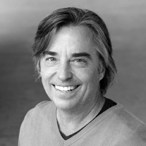 Smiling middle-aged man with gray hair wearing a light sweater against a gray backdrop.